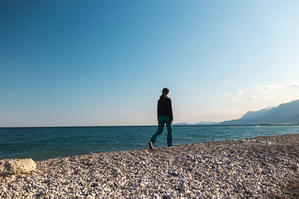 Девушка гуляет по пляжу одна, женщина смотрит на море, брюнетка на фоне моря и гор, отдыхает на морском побережье - Фото, изображение