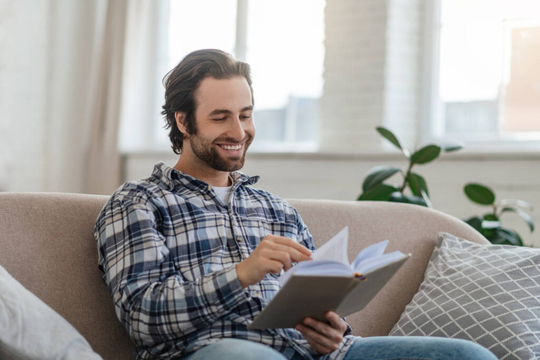 Happy millennial handsome caucasian guy with stubble sits on sofa reads book in living room interior - Photo, image