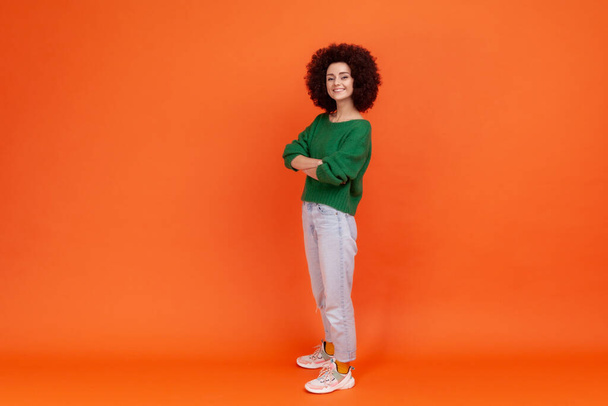Full length of happy confident woman with Afro hairstyle wearing green casual style sweater standing with hands on hips, expressing positive emotions. Indoor studio shot isolated on orange background. - Foto, Imagen