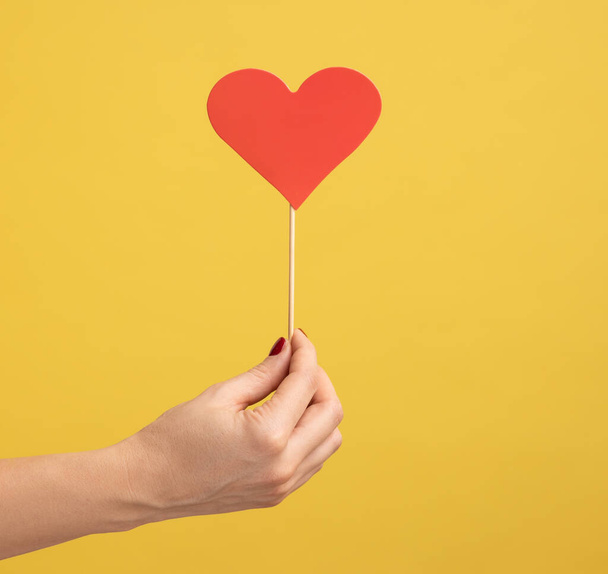 Profile side view closeup of woman hand holding small red heart stick in hand, symbol of love and gentle. Indoor studio shot isolated on yellow background. - Foto, Bild