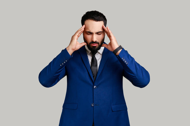 Bearded man frowning and clasping sore head, suffering intense headache, having unbearable migraine, fever and flu symptoms, wearing official style suit. Indoor studio shot isolated on gray background - Photo, Image