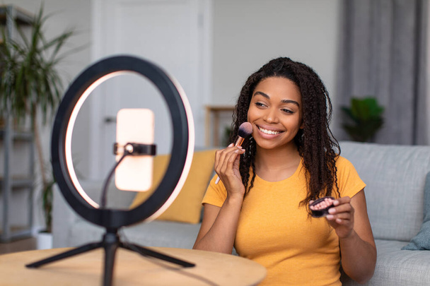 Beauty blogging concept. Happy black lady recording makeup review, using ring lamp and cellphone - Foto, afbeelding