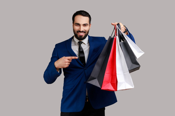 Bearded man pointing finger at paper bags in her hand, pleased with shopping, low prices good quality, wearing official style suit. Indoor studio shot isolated on gray background. - 写真・画像