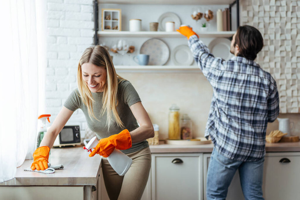 Lächelnde europäische Millennial-Blondine mit Gummihandschuhen und Mann wischt Staub auf hellem Kücheninnenraum - Foto, Bild