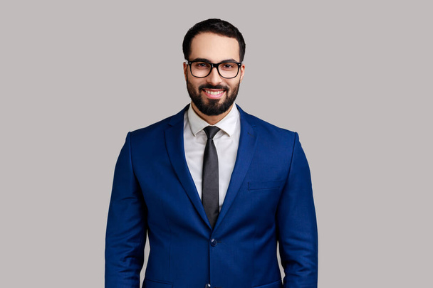 Portrait of happy smiling satisfied bearded businessman in eyeglasses, looking at camera with positive expression, wearing official style suit. Indoor studio shot isolated on gray background. - Photo, image