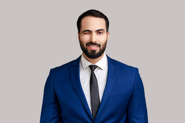 Portrait of bearded businessman in good mood, smiling broadly and winking at camera with toothy smile, wearing official style suit. Indoor studio shot isolated on gray background. - Photo, image