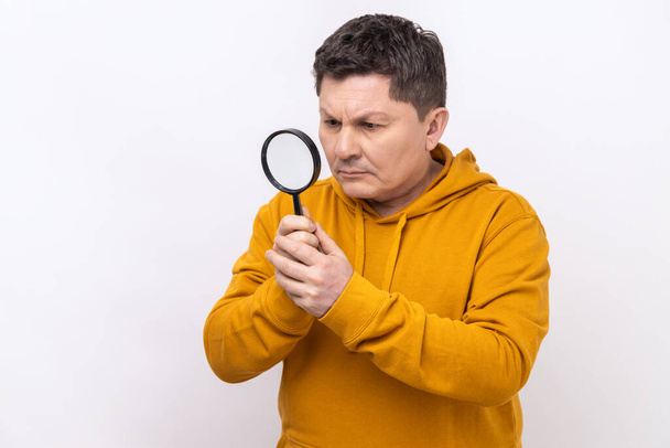 Middle aged man looking through magnifying glass, spying, finding out something, exploring crime scene, inspecting, wearing urban style hoodie. Indoor studio shot isolated on white background. - Foto, Bild