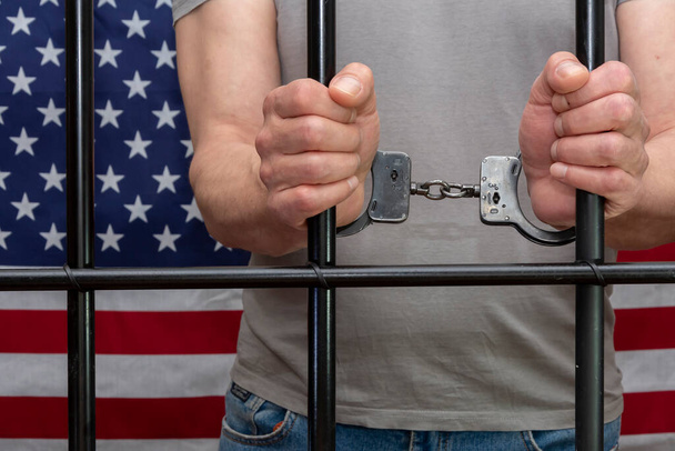 Um homem está algemado numa cela atrás das grades no fundo de uma bandeira americana. Conceito: o prisioneiro no tribunal, o veredicto do tribunal para a pessoa condenada, pena de prisão, lei. - Foto, Imagem