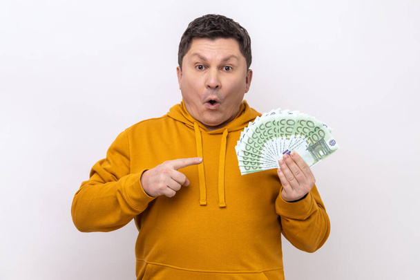 Portrait of amazed shocked man holding pointing at many euro banknotes looking at camera with open mouth, winning lottery, wearing urban style hoodie. Indoor studio shot isolated on white background. - Photo, image