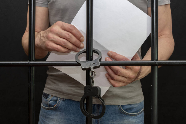 Ένας άντρας με χειροπέδες κρατάει ένα λευκό χαρτί στα χέρια του πίσω από τα κάγκελα ενός κελιού. Έννοια: μια αίτηση για επιείκεια, η τελευταία λέξη, ένας κρατούμενος στο δικαστήριο, μια δικαστική απόφαση σε ένα καταδικασμένο πρόσωπο - Φωτογραφία, εικόνα