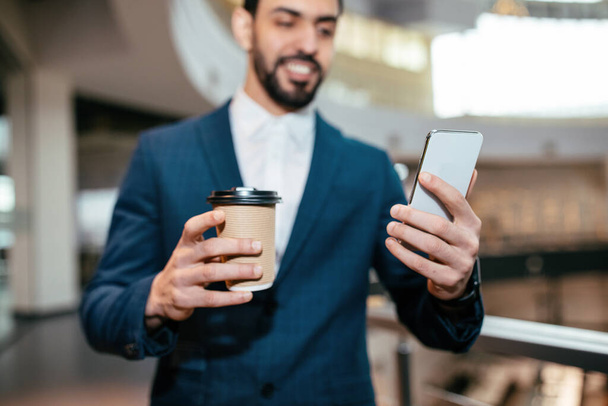 Felice uomo d'affari musulmano millenario con la barba in giacca e cravatta con tazza di caffè guarda smartphone in ufficio interno - Foto, immagini