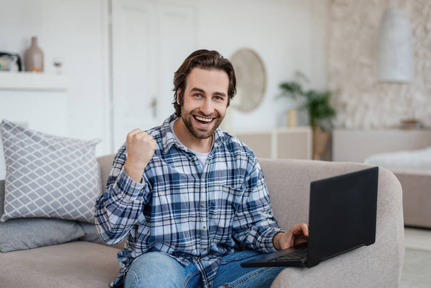 Glad young caucasian man with stubble sits on sofa make victory gesture, sits on sofa with laptop in living room - Фото, изображение