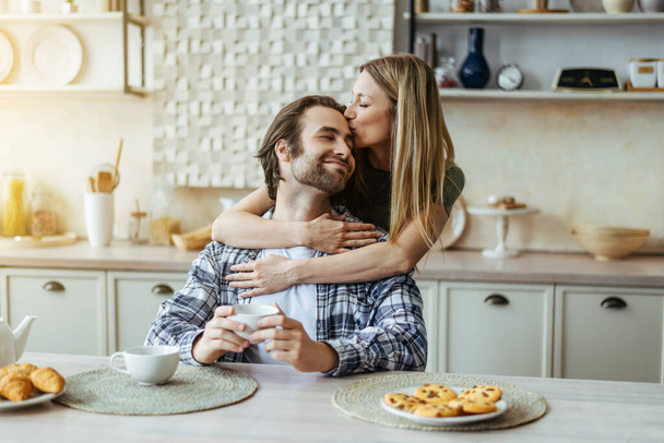Happy caucasian millennial blonde wife kissing man with stubble on light kitchen interior, sun flare - Foto, Imagen