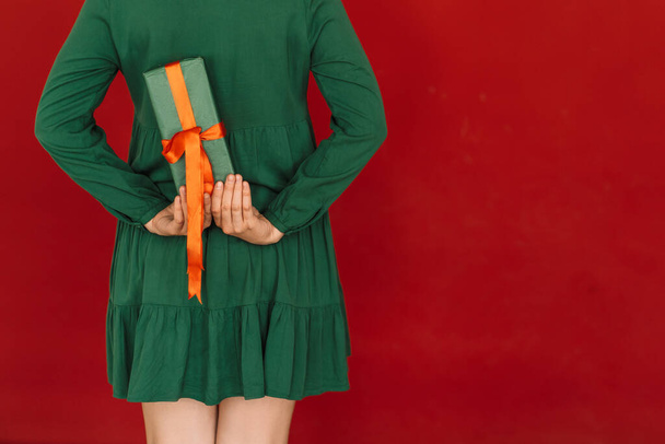 Chica en vestido verde sostiene regalo detrás de ella contra fondo rojo - Foto, imagen