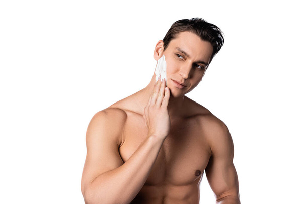 brunetka bez trička muž nanášení pěny na holení a odvrací pohled izolované na bílém - Fotografie, Obrázek