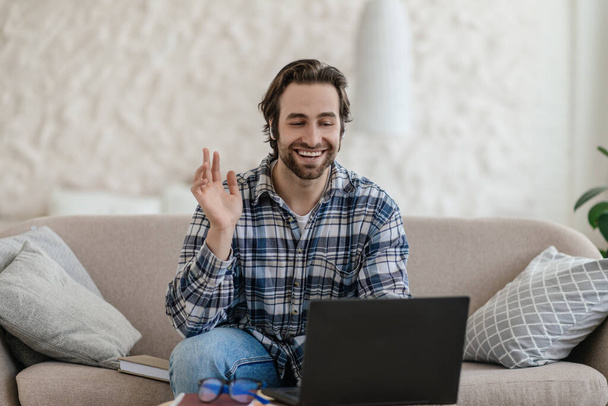 Glad millennial caucasian guy with stubble sits on sofa waving hand and looking at laptop in living room - Foto, imagen