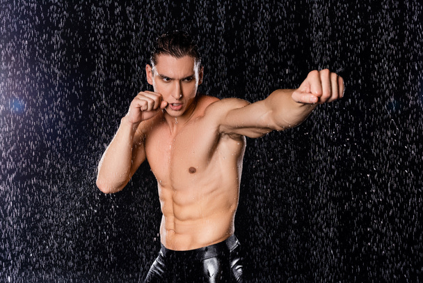 hombre fuerte con torso muscular en pose de boxeo bajo la lluvia sobre fondo negro - Foto, imagen
