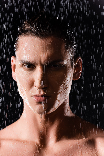 retrato del hombre mojado mirando a la cámara bajo gotas de agua cayendo sobre fondo negro - Foto, Imagen