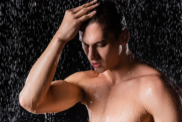 man with closed eyes touching hair while showering on black background - Photo, Image
