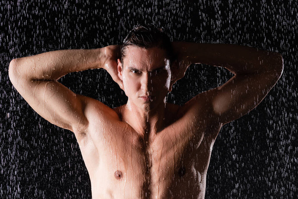 hombre mojado posando con las manos detrás de la cabeza bajo gotas de agua sobre fondo negro - Foto, Imagen