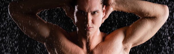 muscular man posing with hands behind head under shower on black background, banner - Photo, Image