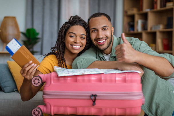 Vacation concept. Excited african american spouses holding passports and showing thumb up, leaning on suitcase - Foto, Imagen