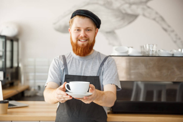 Coffee Business owner Concept - Portrait of happy young bearded caucasian barista in apron with confident looking servicing hot coffee to customer in coffee shop counter. - Photo, Image