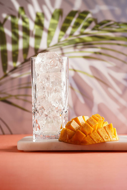 copa de cóctel alta llena de cubitos de hielo desigual y cortar en cubos de mango medio sobre fondo de palmera tropical, efecto máscara de gobo - Foto, Imagen