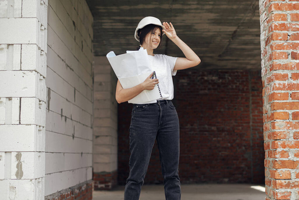 Jonge vrouwelijke ingenieur of bouwvakker in harde hoed glimlachend tegen de bouw van nieuwe moderne huis. Stijlvolle vrolijke vrouwelijke architect met blauwdrukken en plannen op de bouwplaats - Foto, afbeelding