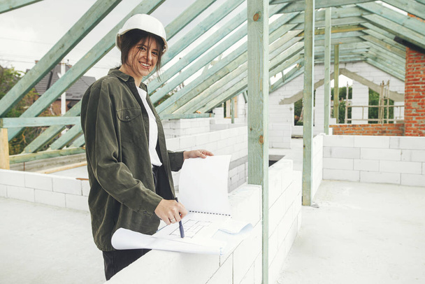 Élégant architecte femme heureuse vérifier les plans contre charpente de toit en bois de ferme moderne. Jeune ingénieure ou ouvrière de la construction en casque rigide avec des plans sur le chantier - Photo, image