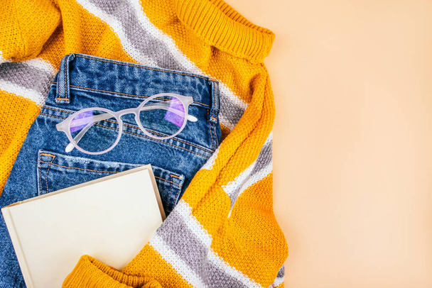 Trendy herfstmode flatlay met gele trui, blauwe jeans, witte brillen en boek. - Foto, afbeelding