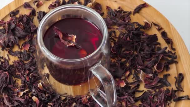 Tè rosso in una tazza di vetro su sfondo bianco. Il tè di ibisco rosso viene versato in un vetro trasparente con doppie pareti. Una tazza di tè rotante su una tavola di legno. - Filmati, video