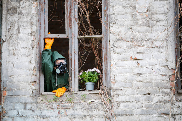 A woman in a raincoat and gas mask collects a flower from a scorched, poisonous earth. Air pollution concept. Ecological catastrophy. - Foto, imagen
