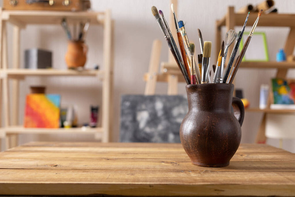 Artist supplies at wooden table. Paint brush in clay jug and art painter tools on wood desk background texture. Paintbrush for painting in artist studio workplace - Fotografie, Obrázek
