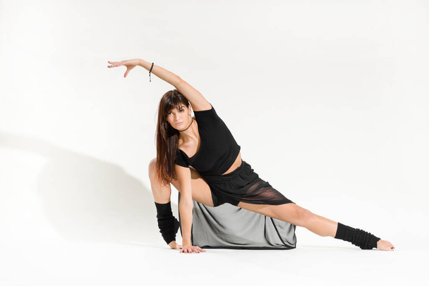 Πλήρες σώμα νεαρής χορεύτριας με μαύρη στολή που εκτελεί πρόσθιο τέντωμα με λυγισμένο σκαλοπάτι και υψωμένο χέρι σε λευκό φόντο - Φωτογραφία, εικόνα