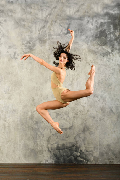 Bailarina ágil de cuerpo completo en maillot levantando brazos y saltando alto mientras baila contra la pared gris grumosa. Concepto de danza. - Foto, imagen