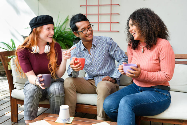 Gruppe von multirassischen Freunden mit Tassen Kaffee plaudern und einander anschauen, während sie auf einem bequemen Sofa im hellen Innenhof sitzen - Foto, Bild