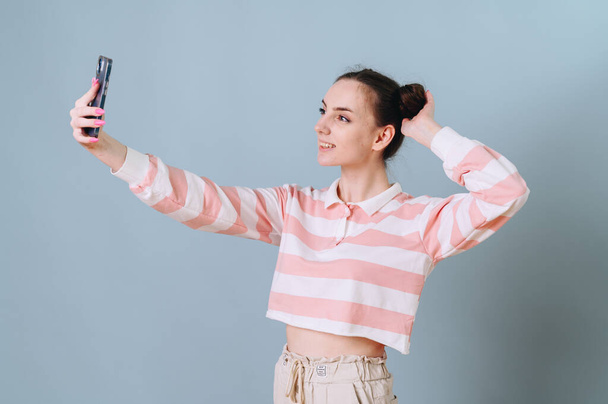 Krásná dívka v pruhovaném svetru vezme selfie na modré pozadí a housky na hlavě. Koncept přátelství, komunikace, virtuální lásky - Fotografie, Obrázek