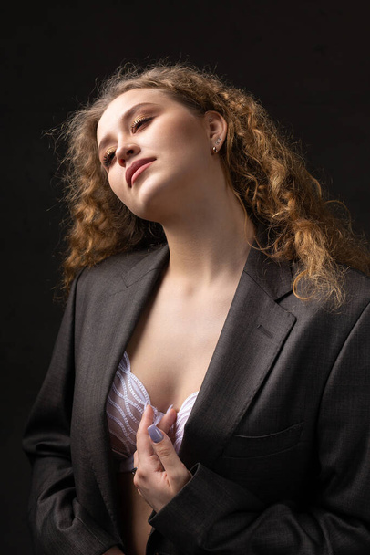 Porträt eines jungen rothaarigen, lockigen sexy Mädchens in einer Jacke mit langen Haaren im Studio. Zeigt ihre kurvenreichen Brüste in einem BH. - Foto, Bild