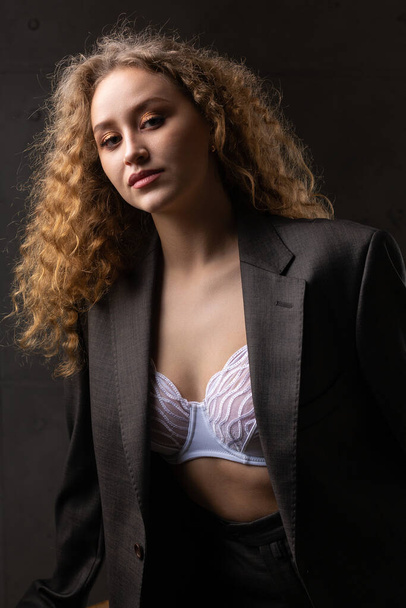 Portrait of a young red-haired curly sexy girl in a jacket with long hair in the studio. Shows off her curvaceous breasts in a bra. - Foto, Imagen