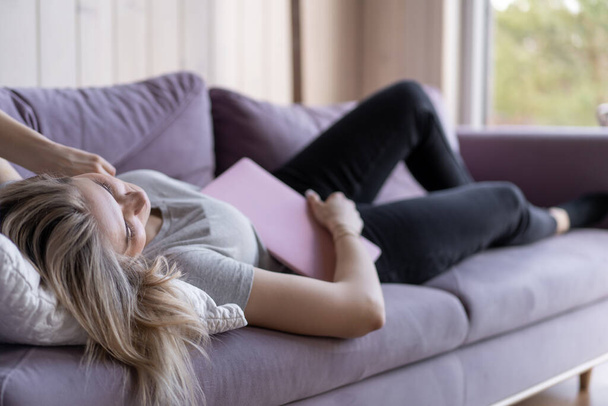 Young blonde in pink T-shirt and jeans fell asleep at home on couch with laptop in her hands. Work at home online. Weekend. Sleep problems. Overwork. Burnout. - Фото, изображение