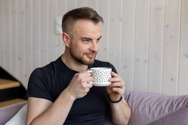 Glücklicher junger Mann in schwarzem T-Shirt sitzt auf gemütlichem Sofa und genießt heißen Tee. Ruhe und Entspannung. Wohnkomfort. Glückliche Momente. Positive Emotionen. - Foto, Bild