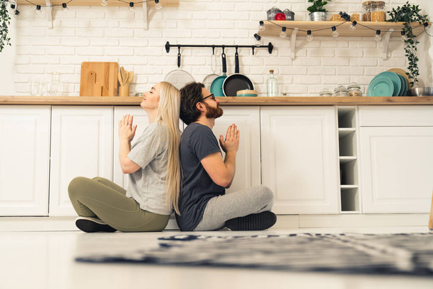 Young adult couple sitting on the floor back to back meditating together in a cosy well-equipped kitchen. Indoor shot - Zdjęcie, obraz