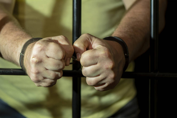 Ένας άντρας με χειροπέδες σε ένα κελί πίσω από τα κάγκελα. Έννοια: φυλακισμένος σε δικαστήριο, δικαστική απόφαση σε καταδικασθέντα, ποινή φυλάκισης. - Φωτογραφία, εικόνα