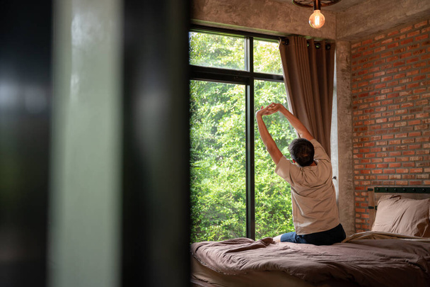 A lazy man happy waking up in the bed rising hands to window in the morning with fresh feeling relax in a brown room made of bricks. - Photo, image
