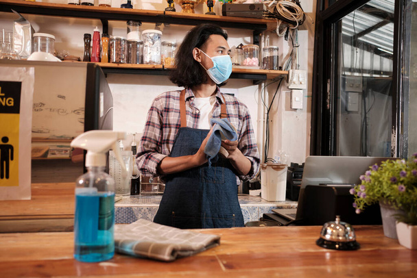 Alcohol sanitizer for healthy protection in a cafe, Asian male barista in face mask use laptop, waiting for coffee order in COVID19 pandemic quarantine, the economic impact for small business startup. - Foto, Imagen