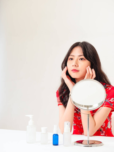 Portrait of a young asian woman in traditional cheongsam qipao dress looks at mirror and does skin care isolated on white background. - Φωτογραφία, εικόνα
