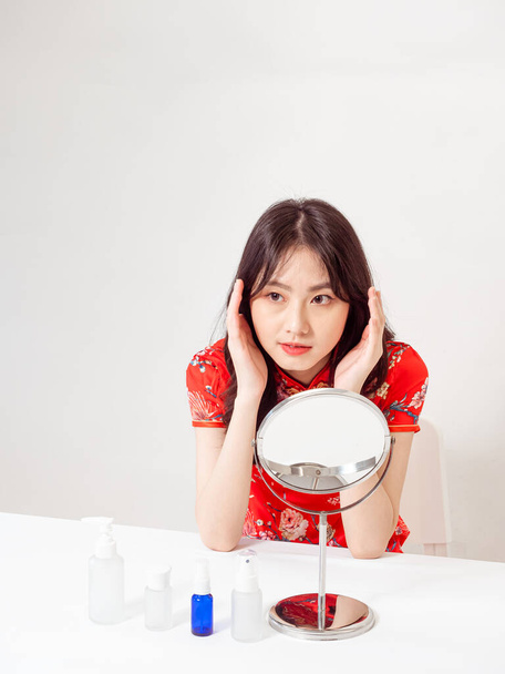 Portrait of a young asian woman in traditional cheongsam qipao dress looks at mirror and does skin care isolated on white background. - Φωτογραφία, εικόνα