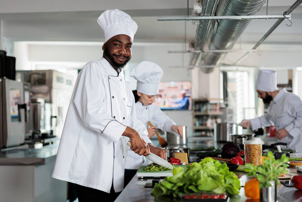 Experto en gastronomía afroamericana sonriendo con confianza mientras corta verduras frescas para la cena y posa para la cámara - Foto, imagen