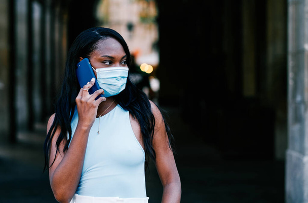 Young woman with a protective mask talking on the phone while standing outdoors. - 写真・画像